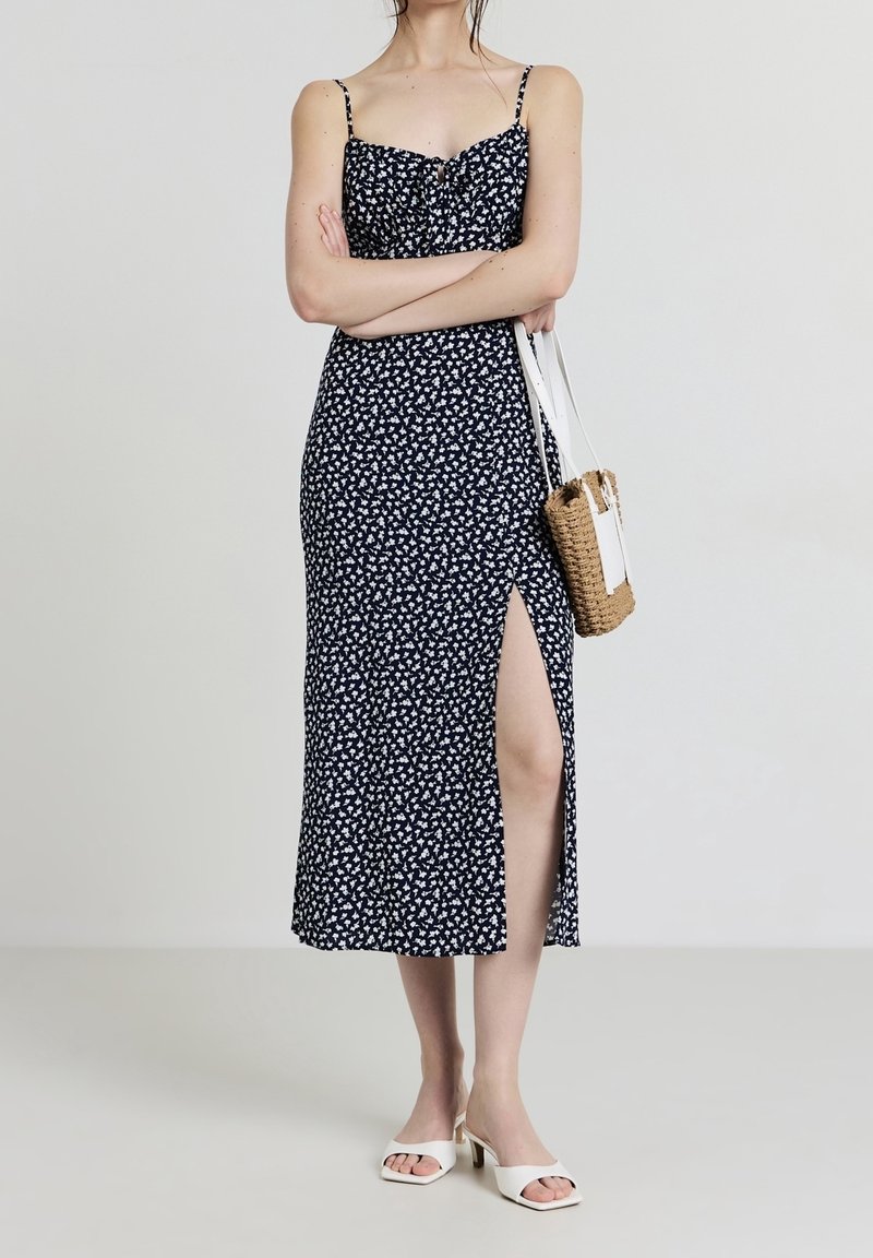 Femme portant une robe fleurie noire et blanche à bretelles spaghetti et fente haute sur la cuisse, tenant un sac à main tissé, portant des sandales blanches à talons.
