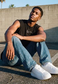 Ung mann i svart ermeløs skjorte og blå jeans sitter på asfalt med armene hvilende på knærne mot en grå betongblokkvegg.