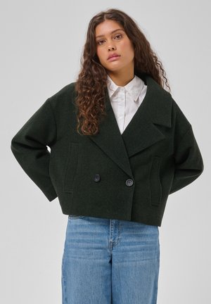 Vrouw met lang krullend haar draagt een donkergroene double-breasted blazer, een wit overhemd met kraag en blauwe jeans, staand tegen een effen achtergrond.