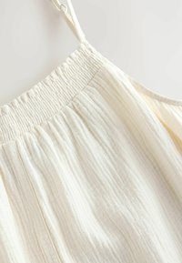 Haut en tissu léger couleur crème avec encolure smockée élastique et bretelles ajustables ; possède une surface douce et texturée avec des lignes verticales.