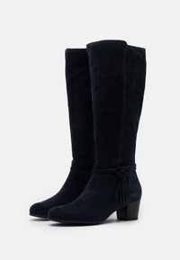 Knähöga stövlar i marinblå mocka med spetsig tå och blockklack. Har en knutdetalj runt vristen och kontrasterande sömmar längs sömmarna.