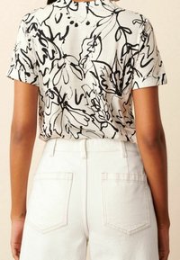 Camisa de manga corta con base blanca y diseño floral negro. Presenta mangas dobladas y un ajuste holgado, combinada con pantalones de color claro.