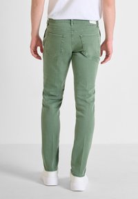 Vaqueros de mezclilla verde con un ajuste slim, diseño de cinco bolsillos y costuras en contraste, combinados con zapatillas blancas. Textura suave, sin patrones visibles.