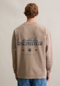 Beżowa bluza z luźnym krojem, okrągłym dekoltem i ściągaczem na dole. Na plecach znajduje się niebieski napis i logo z napisem "stockholm ATHLETES CLUB".