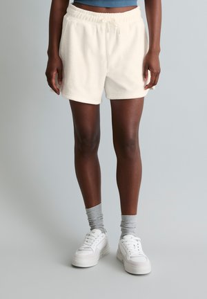 Persoon die crème-kleurige casual shorts, grijze sokken en witte sneakers draagt, staand tegen een effen lichte achtergrond.