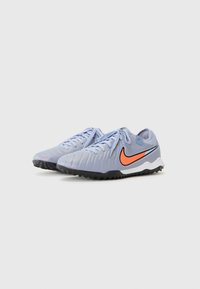 Nike sportssko med en lysegrå overdel, tekstureret overflade, orange swoosh-logo, og sort gummisul for trækkraft.