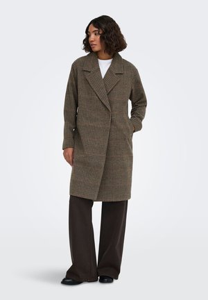Femme aux cheveux courts et bouclés portant un long manteau marron à carreaux, un pantalon large foncé, des chaussures noires et une chemise blanche, debout les mains dans les poches.