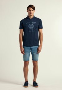 Camisa polo de manga corta en color marino con un diseño gráfico, combinada con pantalones cortos de mezclilla claros y zapatillas deportivas oscuras con suela de goma.