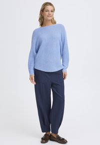 Femme debout portant un pull en tricot bleu, un pantalon large bleu marine et des mocassins marron, sur un fond blanc uni.