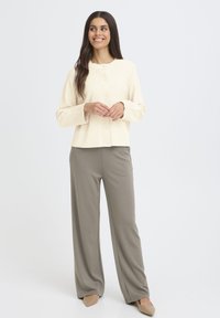 Kremo spalvos mygtukinė kardiganas su raukšlėtu tekstūra, derinamas su šviesiai pilkomis plačiomis kelnėmis. Modelis stovi su rankomis sukryžiuotomis.