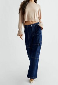 Nainen, jolla on päällään beige lyhyt neulepusero, tummansiniset leveät cargo-farkut ja valkoiset kärjet korkokengät, joissa on kultaiset yksityiskohdat.