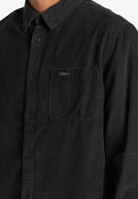 Camisa de pana negra con cuello abotonado, bolsillo frontal y textura de rayas horizontales. Presenta seis botones para el cierre.