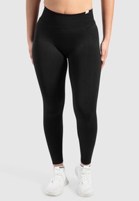 Leggings noirs taille haute en tissu extensible avec une texture lisse. Design ajusté, longueur cheville, branding minimal sur la taille.