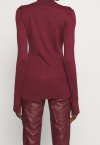 Suéter de punto burdeos con cuello alto y mangas largas, con ribete en el dobladillo y puños. Combinado con pantalones de cuero burdeos a juego.
