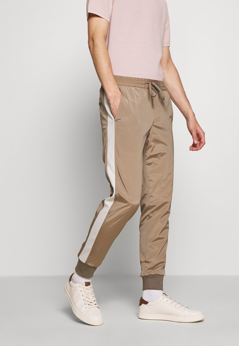 Pantalones jogger beige hechos de material ligero, con una franja lateral blanca, cintura elástica y puños acanalados. Usados con zapatillas blancas.