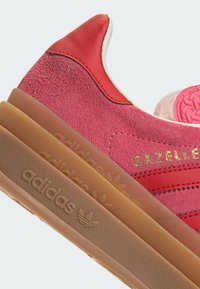 Sapatilhas em camurça e couro vermelho com uma sola de borracha texturizada, apresentando a marca "adidas" em relevo e o logótipo dourado "GAZELLE".