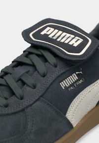 Teniska Puma Palermo v námorníckej modrej semišovej farbe s tmavozelenými šnúrkami a bielym koženým pruhom. Obsahuje výraznú čierno-bielu logo značku.
