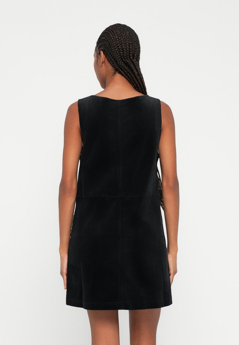 Femme aux longs cheveux tressés portant une robe noire en velours côtelé sans manches, debout de dos devant un fond clair uni.