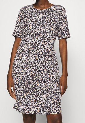 Frau trägt ein kurzärmeliges, knielanges Kleid mit einem dicht gemusterten schwarzen, weißen und orangen Blumenmuster auf hellem Hintergrund.
