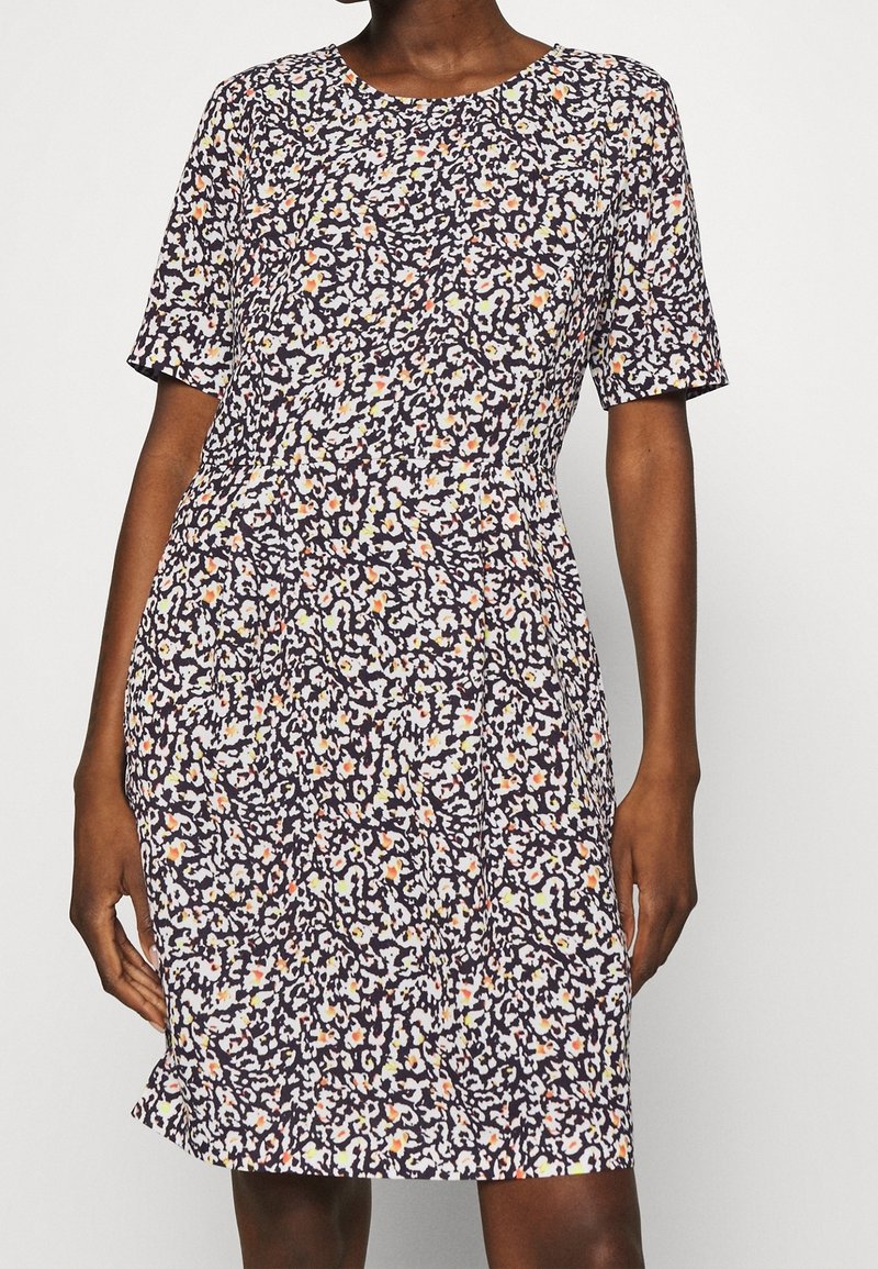 Femme portant une robe à manches courtes, longueur genoux, avec un motif floral dense noir, blanc et orange sur fond clair.