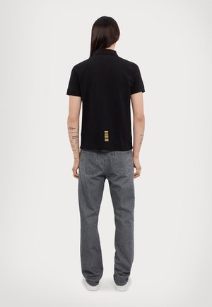 Person mit langen Haaren, der ein schwarzes kurzärmeliges Hemd und graue Jeans trägt und mit dem Rücken zu einem schlichten Hintergrund steht.