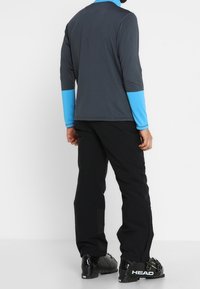 Camisa de manga larga de color gris oscuro con puños azules brillantes, combinada con pantalones negros y botas de esquí negras con detalles verdes.