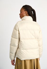 Jachetă puffer din bej deschis, cu un design matlasat, guler înalt și manșete elastice. Material moale și neted, cu o textură ușor lucioasă.