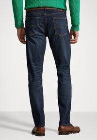 Mörkblå denimjeans med smal passform, med lätt viskning, två bakfickor och bruna läderskor. Stickad grön tröja syns.