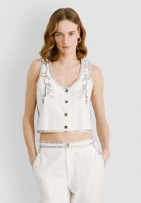 Bijela skraćena majica s V-izrezom, ukrašena dekorativnim detaljima srebrne boje i zatvaranjem na gumbe sprijeda.