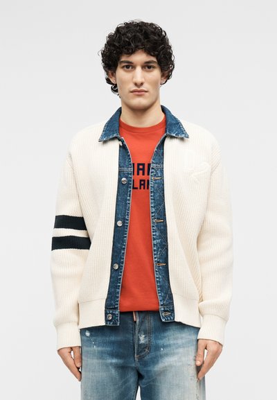 Jeune homme aux cheveux bouclés portant un cardigan en maille blanc cassé à rayures noires, superposé sur une veste en denim et un t-shirt rouge à motif, jean bleu.