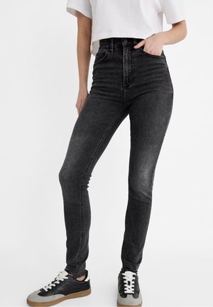 Personne portant un jean skinny noir taille haute et des baskets noires et grises avec des lacets blancs, une main dans la poche, sur un fond uni.