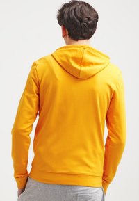 Sudadera con capucha amarilla de textura suave y lisa, con capucha ajustable mediante cordón y mangas largas, mostrada desde atrás junto con pantalones grises.