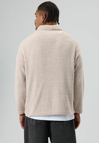 Tankleurige, pluizige trui met kraag, zachte textuur, lange mouwen en een comfortabele pasvorm. Draagt over een witte laag met donkere broek.