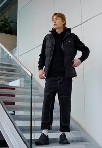 Gilet puffa nero sopra un felpa nera con cappuccio, abbinato a pantaloni neri con risvolto caratterizzati da cuciture bianche. Il modello è in piedi su una scala.