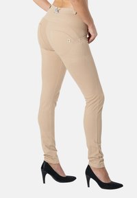 Nainen pukeutuneena beigeihin istuviin housuihin, joissa on takataskut, ja mustiin korkokenkiin, seisomassa yksiväristä taustaa vasten.