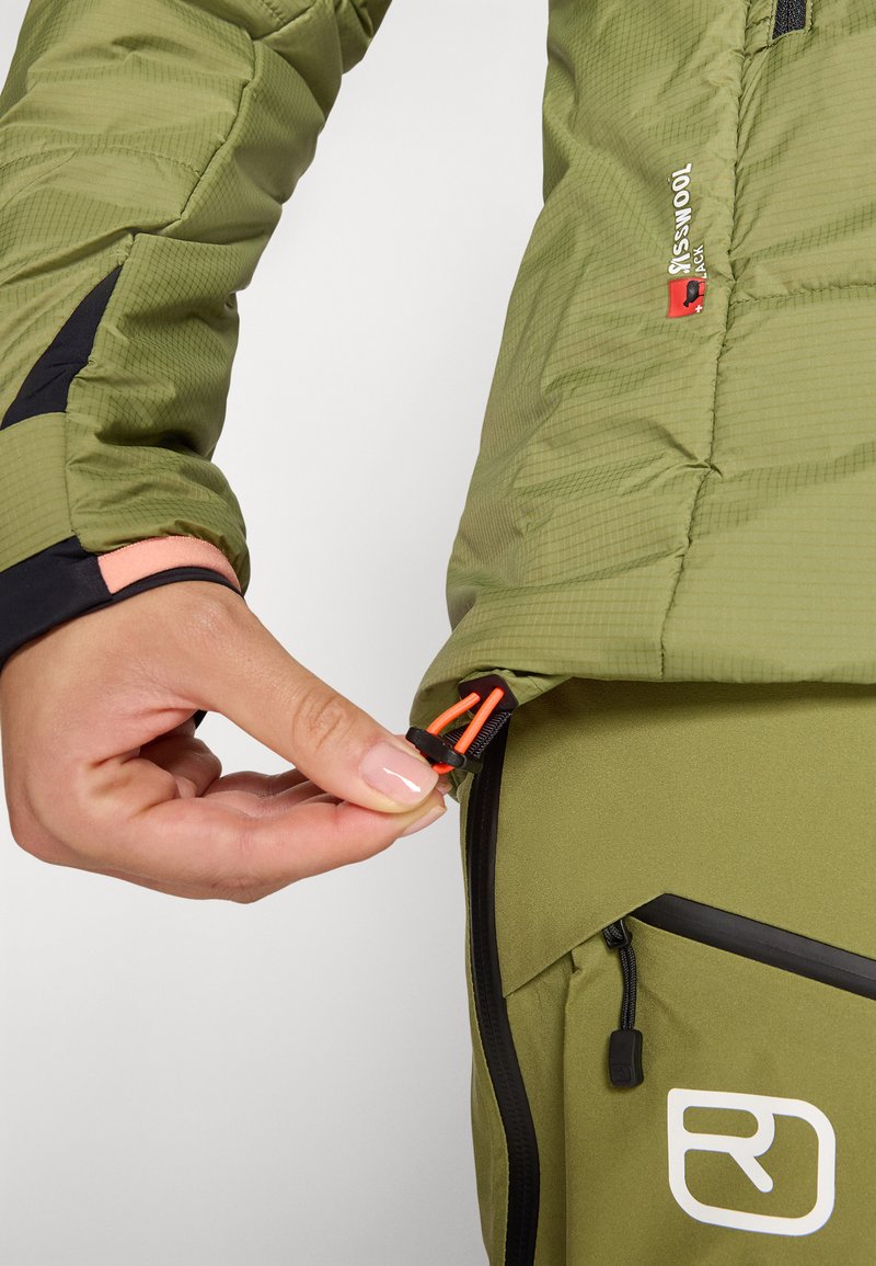 Grüne isolierte Jacke mit einer Ripstop-Oberfläche, die kontrastierende schwarze und pinke Akzente aufweist. Die Hand zeigt den verstellbaren Reißverschluss-Zip.