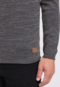 Pull en tricot gris avec une finition texturée, présentant un ourlet cousu et une étiquette en tissu marron sur le côté inférieur, porté avec un pantalon noir.