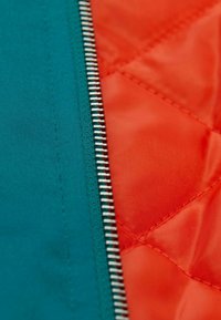 Quilted Jacke mit einem tealfarbigen Außenstoff und einem orangefarbenen Innenfutter, mit einem silbernen Reißverschluss entlang der Naht und einer glatten Textur.