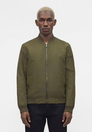 Homme aux cheveux blonds courts portant une veste bomber vert olive zippée et un pantalon noir, debout devant un fond blanc uni.