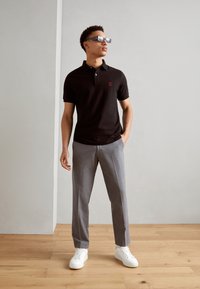 Schwarzes Poloshirt mit einem kleinen roten Logo, graue Hose und weiße Sneakers. Das Model trägt Sonnenbrille mit Reflektion und steht auf einem Holzfußboden.