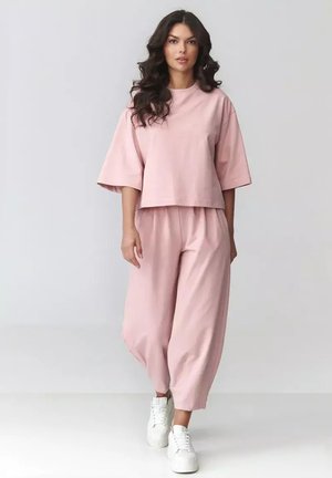 Kobieta idąca do przodu, ubrana w luźny różowy dwuczęściowy strój z szerokimi rękawami i spodniami, w połączeniu z białymi trampkami, na jednolitym tle.