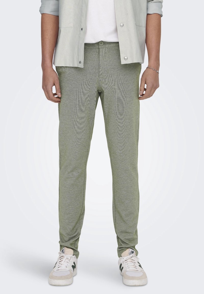 Homme portant un pantalon slim vert, des baskets blanches, une chemise blanche et une veste ouverte gris clair, debout devant un fond blanc.