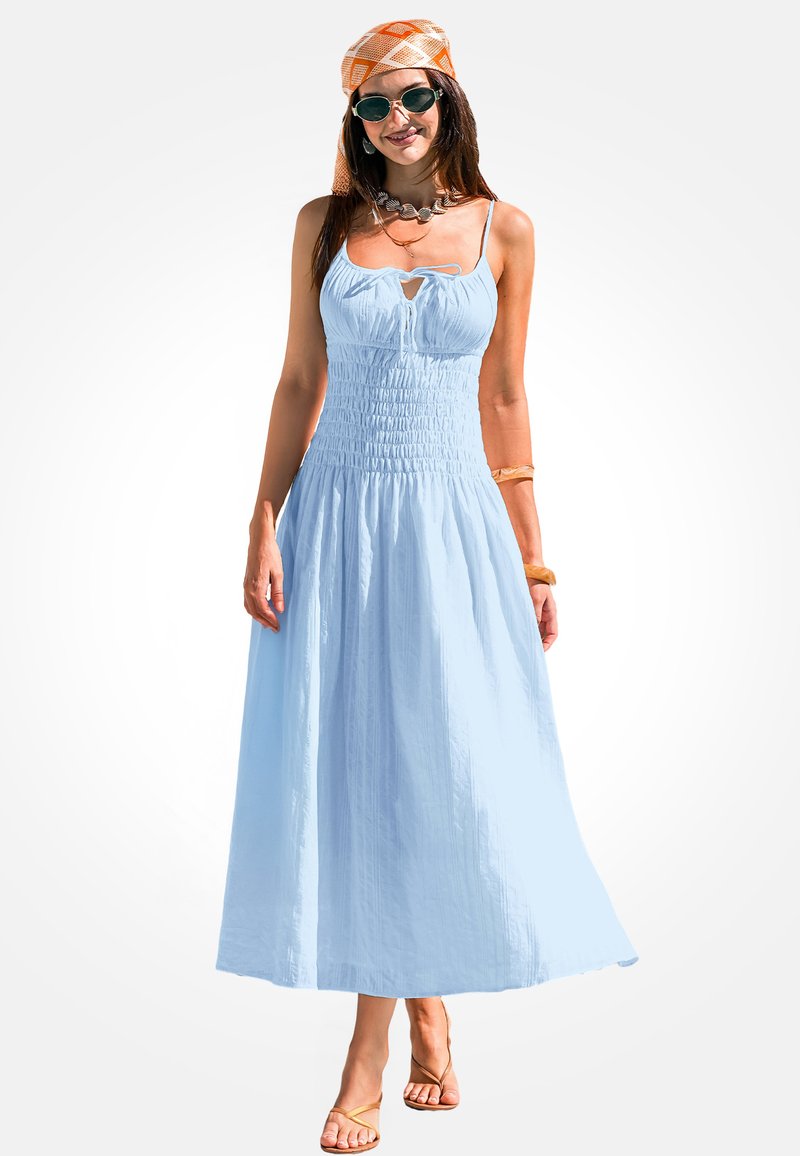 Lichtblauwe maxi-jurk met een smocktop, spaghettibandjes en een sleufdetail. Model draagt sandalen en een oranje met patroon hoofddeksel.