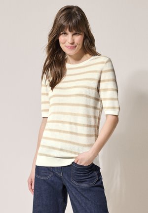 Pull court à manches courtes en crème avec des rayures horizontales beiges. Encolure ronde et coupe décontractée. Porté avec un jean bleu foncé.