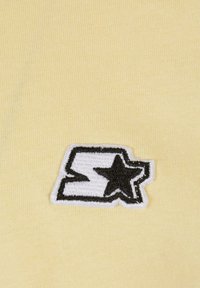 Žltá látka s ryhovanou textúrou obsahuje čierno-biele vyšívané logo, ktoré pozostáva z štýlizovaného písmena a hviezdy.