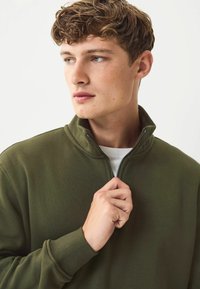 Olivovo zelená mikina na zips s vysokým golierom, vyrobená z mäkkého materiálu. Disponuje textúrovaným povrchom a rebrovanými manžetami.