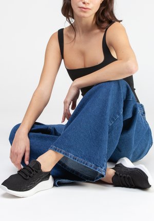 Sneakers in maglia nera con tomaia testurizzata e suole bianche. Indossati con jeans in denim blu a gamba larga. Lo stile è casual e rilassato.