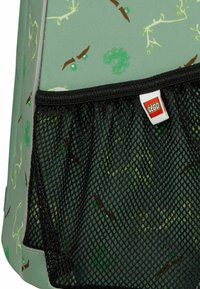 Zeleno blago z rastlinskim vzorcem, črn mrežasti žep in beli obesek z rdečim LEGO logotipom na strani nahrbtnika.