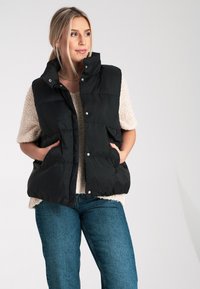 Czarna kamizelka puchowa z wysokim kołnierzem, z zatrzaskami, bocznymi kieszeniami i pikowanym wzorem, noszona na kremowym swetrze i niebieskich dżinsach.