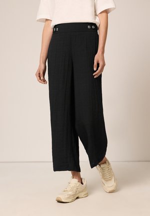 Vrouw draagt zwarte wijde broek met knoopdetails op de tailleband, wit kort mouwtopje en beige sneakers, staand op een lichte vloer.